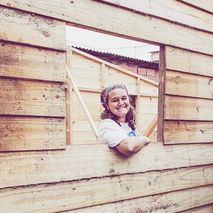 Luciene sorrindo na janela de uma casa de madeira em construção