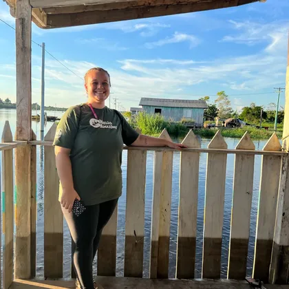 Luciene de pé em uma palafita, com o rio e uma casa de madeira ao fundo