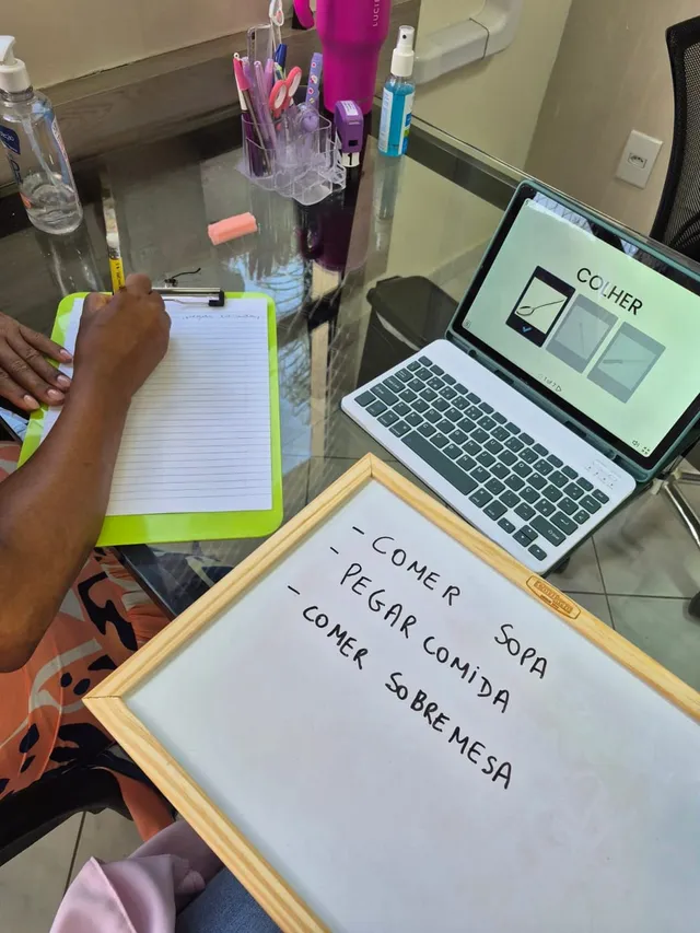 Mesa de terapia com tablet mostrando a palavra COLHER e um quadro com frases escritas